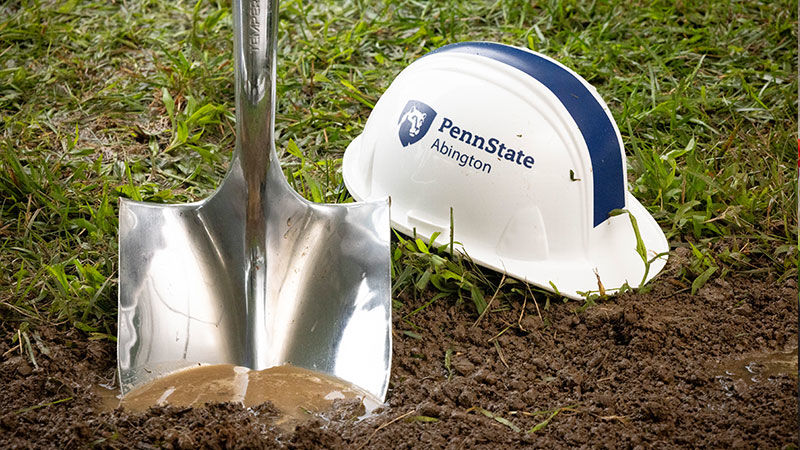 Silver shovel in dirt next to white hard hat with blue letters reading Penn State Abington (near Philadelphia)