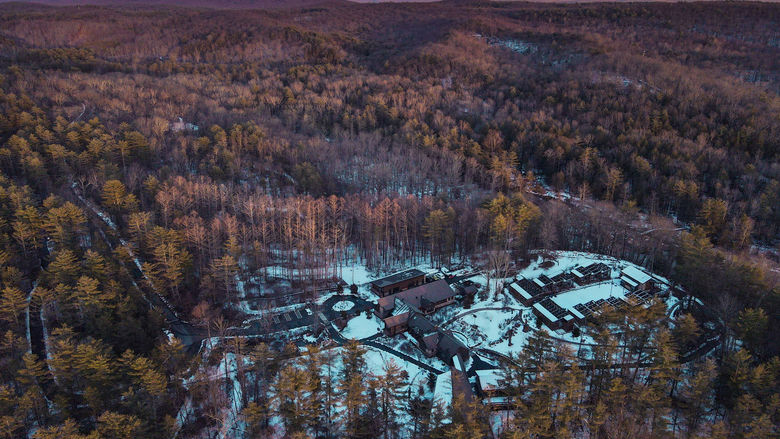 An aerial photograph of a mountainside lined with trees with brown leaves, with a small patch of snowy ground in the center